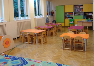 Na fotografii widok sali grupy czerwonej z oknami na wprost z leżącym dywanem i stołami w kolorach fioletowych i pomarańczowych dla dzieci